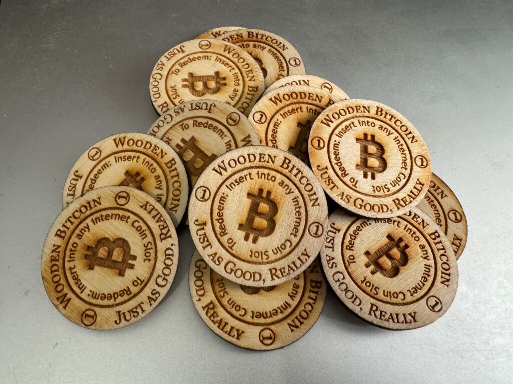 A small pile of wooden disks on a silver background.  An outer circle on each disk reads "Wooden Bitcoin.  Just as Good, Really."  Text on an inner circle reads "To Redeem: Insert into any Internet Coin Slot."  The Bitcoin logo appears at the center of each disk.